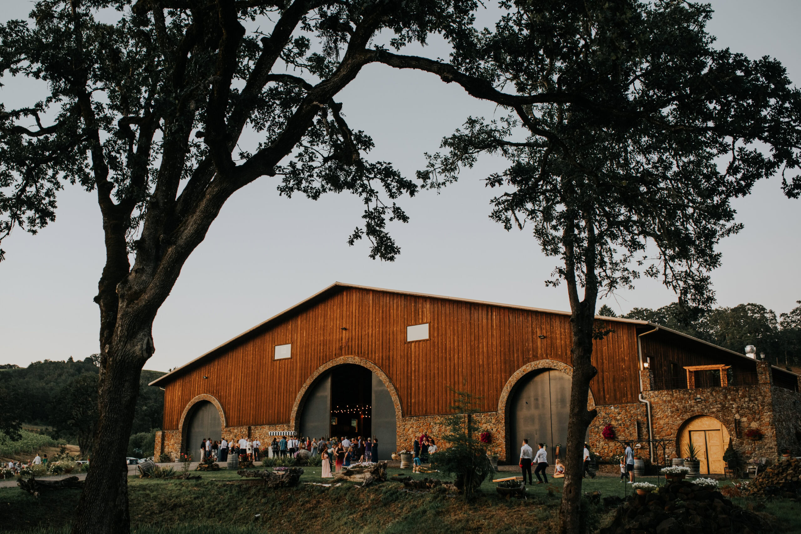 Large wooden barn with arched entrances, surrounded by trees and people gathering outside.