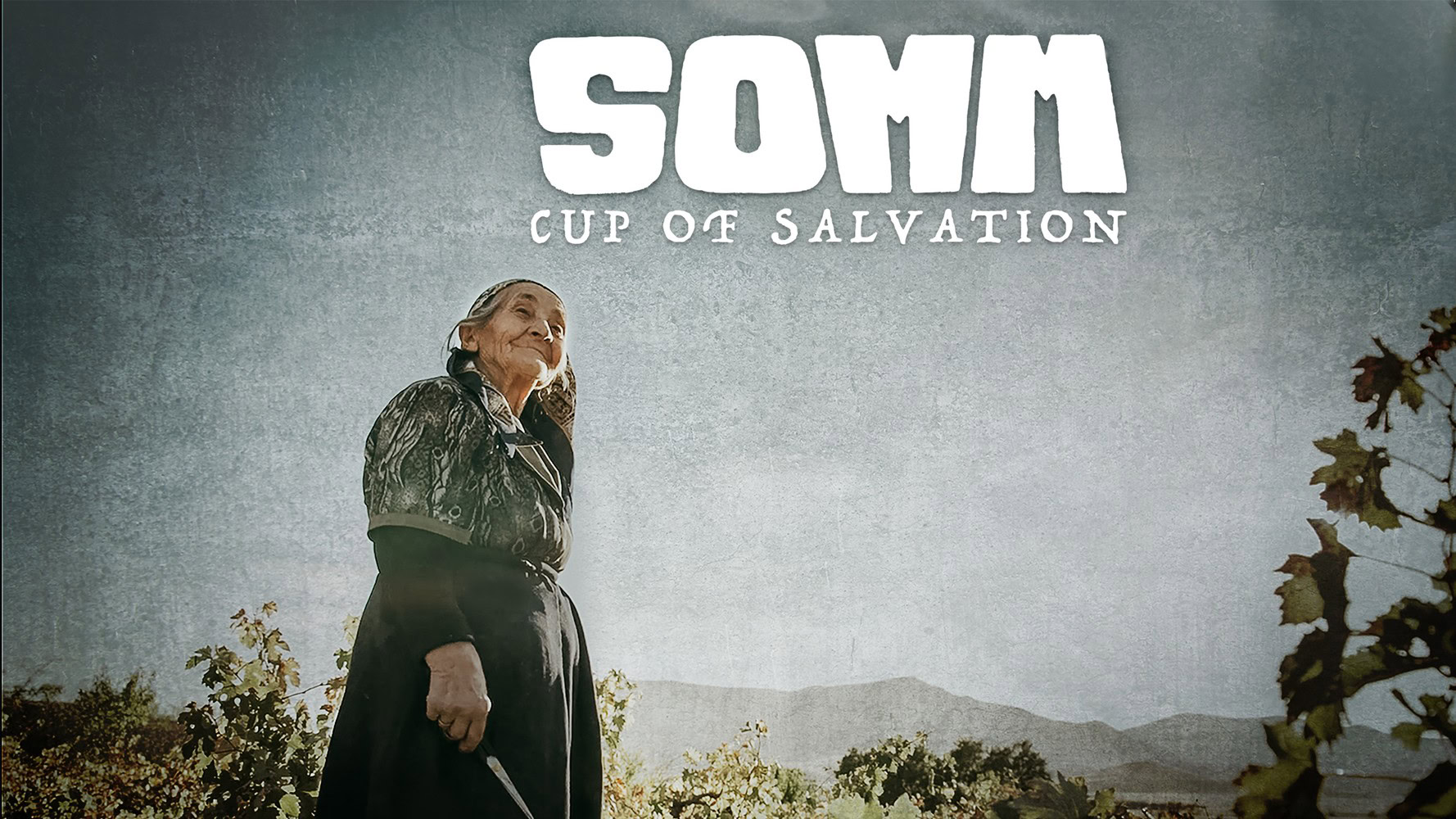 Elderly woman standing outdoors with mountains in the background, smiling and looking up.