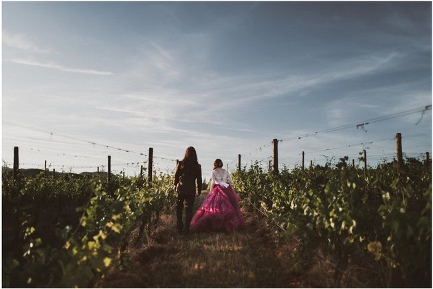 Two women walk through a vineyard under a blue sky with wispy clouds.