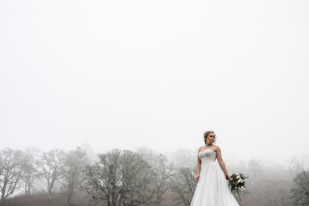Bride in a white wedding dress holding a bouquet outdoors on a foggy day.