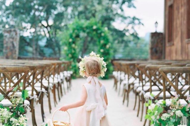 Young girl in a white dress with a floral crown walking down an outdoor aisle at a wedding.