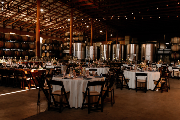 Elegant event setup in a winery with round tables, white tablecloths, and wine barrels in the background.