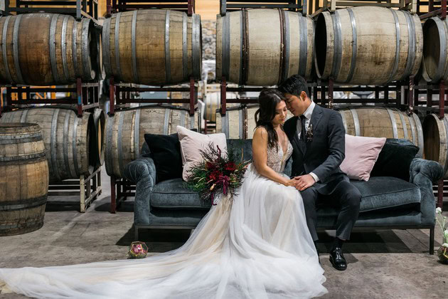 Bride and groom sitting on a sofa in a winery with wine barrels in the background.