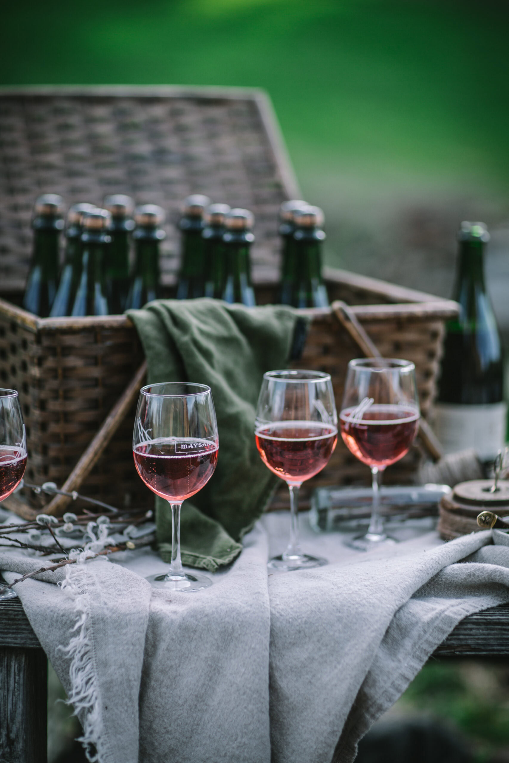 Three glasses of rosé wine on a table outdoors with a basket of wine bottles in the background.